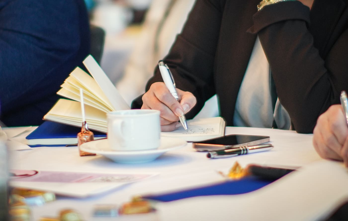Close-up of a hand writing in a small notebook at an event table, beside an open book and a cup of coffee
