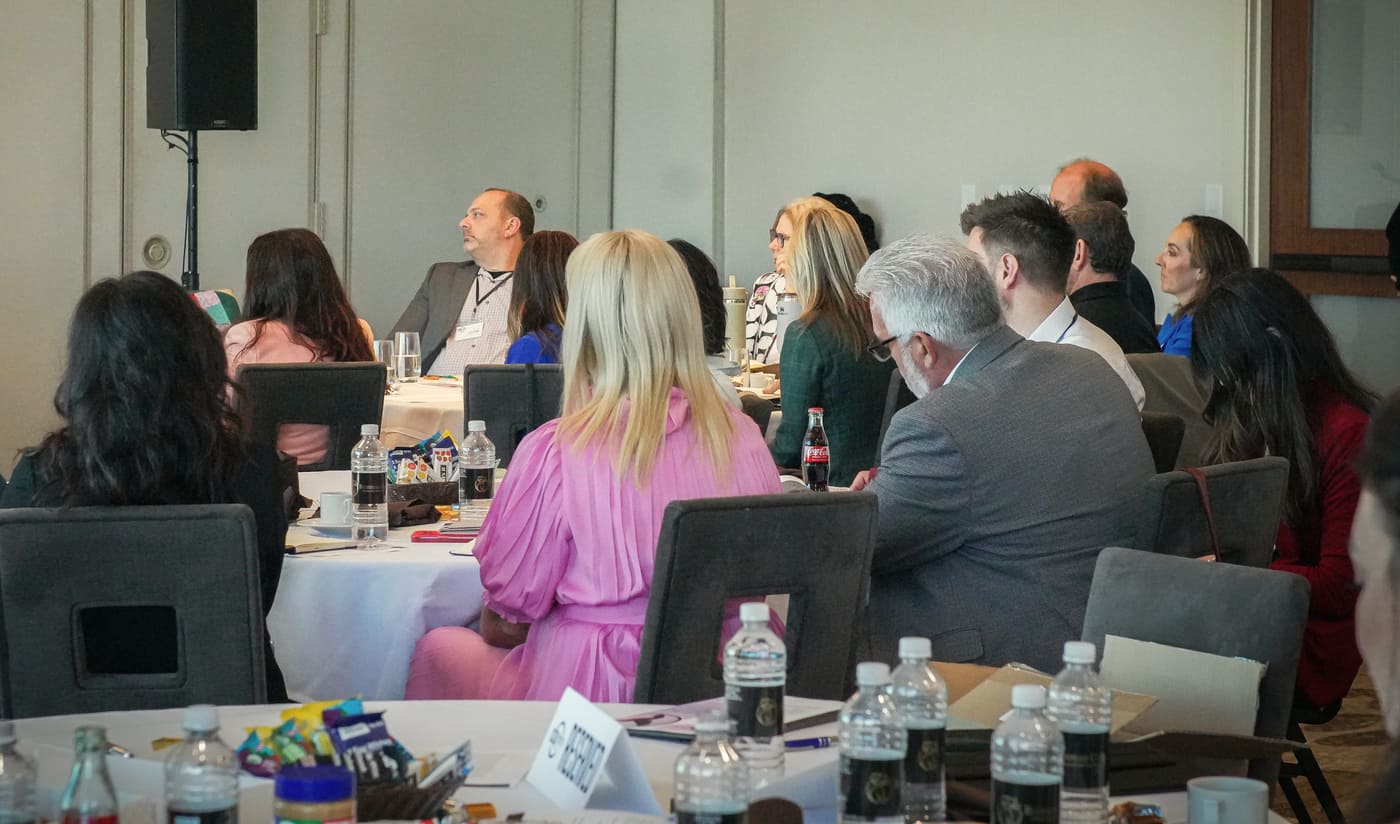 Attendees seated at round tables in a seminar room, seen from behind as they listen
